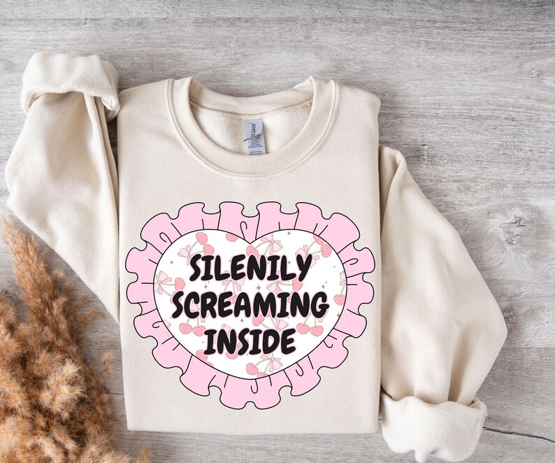 K&ouml;nnte beinhalten: Ein cremefarbener Pullover mit einem rosafarbenen herzf&ouml;rmigen Design. Das Herz hat einen wei&szlig;en Hintergrund mit rosa Kirschen und dem Text "SILENILY SCREAMING INSIDE".