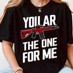 Puede incluir: Camiseta negra con el texto "YOU AR THE ONE FOR ME" y un gráfico de rifle rojo. El gráfico es una ilustración detallada de un rifle con mira telescópica. El texto es en negrita y blanco.