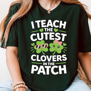 May include: A dark green t-shirt with the slogan "I Teach the Cutest Clovers in the Patch." The design includes two cartoon clovers, one wearing sunglasses and the other a pink bow. The shirt has a crew neck and short sleeves.