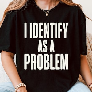 Puede incluir: Camiseta unisex negra con el texto "I IDENTIFY AS A PROBLEM" en letras blancas. La camiseta tiene mangas cortas y cuello redondo. Las palabras están centradas en la parte delantera de la camiseta.