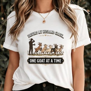 Może przedstawiać: Biała koszulka z napisem "DRIVING MY HUSBAND CRAZY... ONE GOAT AT A TIME." Grafika przedstawia sylwetkę osoby trzymającej kubek i rząd kóz w różnych kolorach.