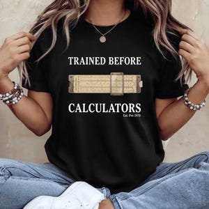 May include: Black t-shirt featuring the text "TRAINED BEFORE CALCULATORS" above a vintage slide rule graphic. The text "Est. Pre-1970" is below the word "CALCULATORS". The shirt is a classic crew neck style.