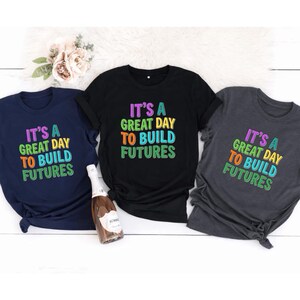 May include: Three t-shirts in navy blue, black, and charcoal gray, each with the text "IT'S A GREAT DAY TO BUILD FUTURES" in colorful, block letters. A bottle of sparkling wine sits in front of the shirts, with a bouquet of flowers in the background.