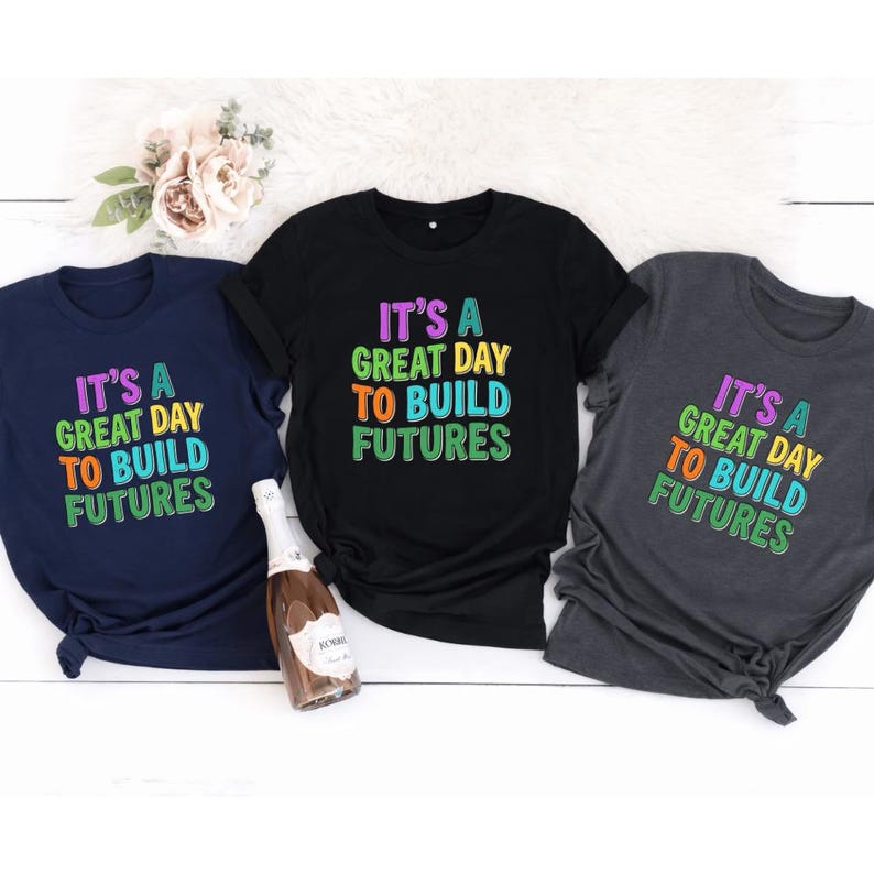 May include: Three t-shirts in navy blue, black, and charcoal gray, each with the text "IT'S A GREAT DAY TO BUILD FUTURES" in colorful, block letters. A bottle of sparkling wine sits in front of the shirts, with a bouquet of flowers in the background.