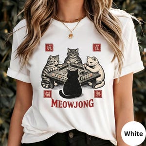 May include: White t-shirt featuring a graphic of four cats playing mahjong around a table. The word "MEOWJONG" is printed below the cats in red. The design includes Chinese characters in red squares.