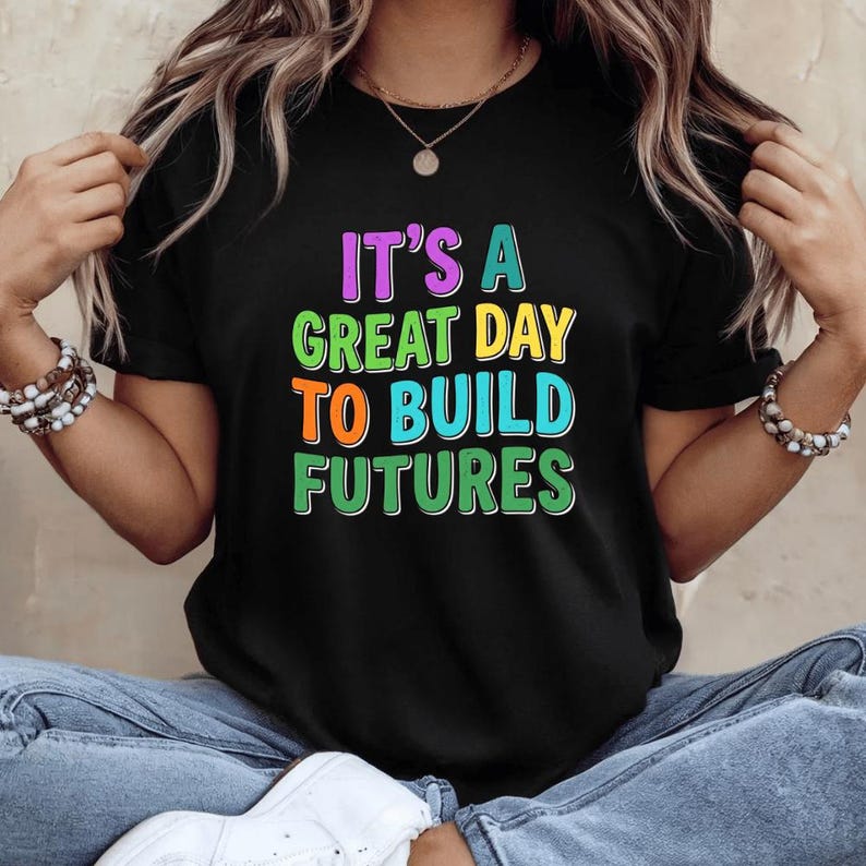 May include: Black t-shirt with the text "IT'S A GREAT DAY TO BUILD FUTURES" in colorful, bold letters. The shirt is a classic crew neck style. The text is in purple, green, orange, and blue.