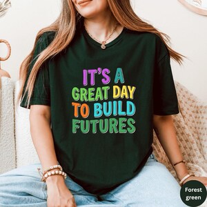 May include: Forest green t-shirt with the text "IT'S A GREAT DAY TO BUILD FUTURES" in colorful, bold letters. The shirt has a crew neck and short sleeves. The text is in purple, green, yellow, orange, and blue.