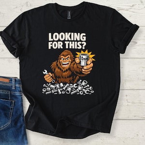 May include: Black t-shirt with the text "LOOKING FOR THIS?" and a cartoon Bigfoot holding a wrench and a 10mm socket. The Bigfoot is surrounded by various nuts and bolts. The shirt is paired with blue jeans.
