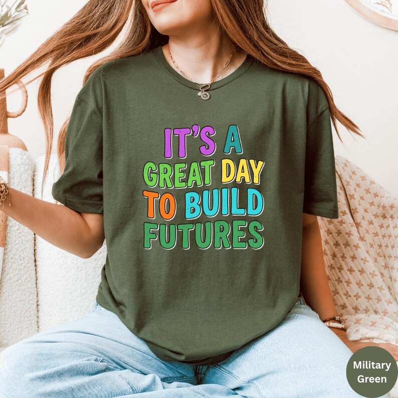 May include: Olive green t-shirt with the text "IT'S A GREAT DAY TO BUILD FUTURES" in colorful, block letters. The shirt is a short-sleeved crew neck style. The model is wearing blue jeans.