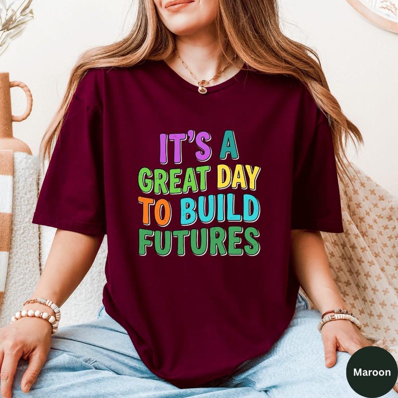 May include: Maroon t-shirt with the text "IT'S A GREAT DAY TO BUILD FUTURES" in colorful block letters. The shirt is short-sleeved and appears to be made of a soft material. The shirt is being worn by a person.