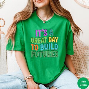 May include: A green t-shirt with the text "IT'S A GREAT DAY TO BUILD FUTURES" in colorful block letters. The shirt is a vibrant green color and has short sleeves. The text is in purple, yellow, orange, and blue.