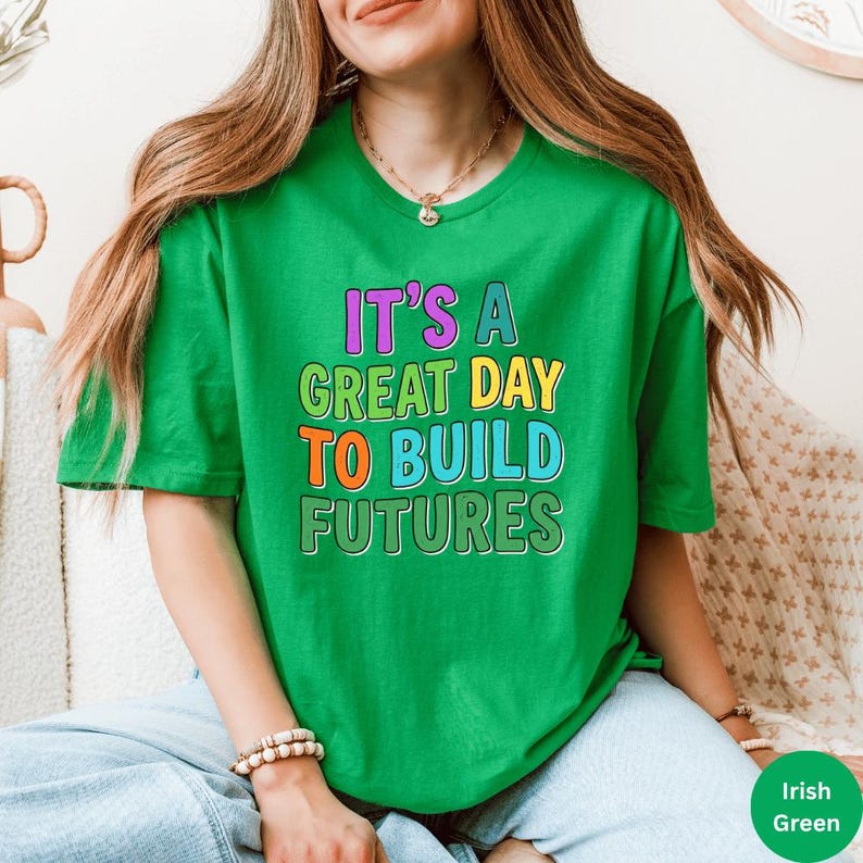May include: A green t-shirt with the text "IT'S A GREAT DAY TO BUILD FUTURES" in colorful block letters. The shirt is a vibrant green color and has short sleeves. The text is in purple, yellow, orange, and blue.