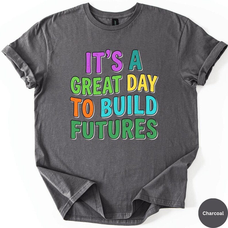 May include: Charcoal-colored t-shirt with the text "IT'S A GREAT DAY TO BUILD FUTURES" in colorful, outlined letters. The shirt has rolled-up sleeves and a casual, relaxed fit. The text is in purple, green, orange, and blue.