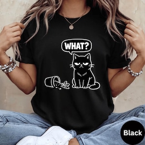 Könnte beinhalten: Schwarzes T-Shirt mit einer weißen Illustration einer mürrischen Katze und dem Wort "WHAT?" in einer Sprechblase. Ein umgestürzter Blumentopf ist neben der Katze abgebildet. Das T-Shirt hat einen Rundhalsausschnitt.