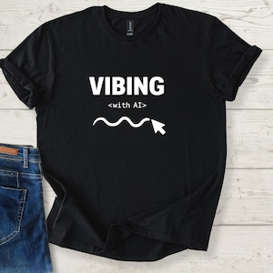 Op de afbeelding: Zwart T-shirt met het woord "VIBING" in witte blokletters, met "<with AI>" eronder. Een witte golvende lijn en een cursorafbeelding staan onder de tekst. Een blauwe jeans is links zichtbaar.