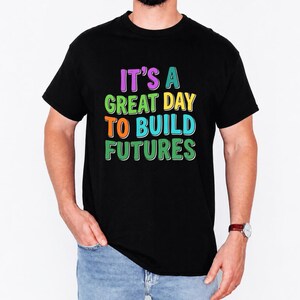 May include: Black t-shirt with the text "IT'S A GREAT DAY TO BUILD FUTURES" in colorful, bold letters. The shirt is a classic crew neck style, suitable for casual wear.