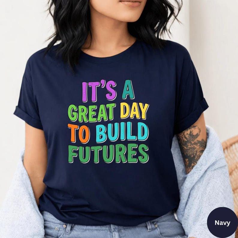 May include: Navy blue t-shirt with the text "IT'S A GREAT DAY TO BUILD FUTURES" in colorful, bold letters. The shirt has short sleeves and a crew neck. The text is in purple, green, orange, and blue.