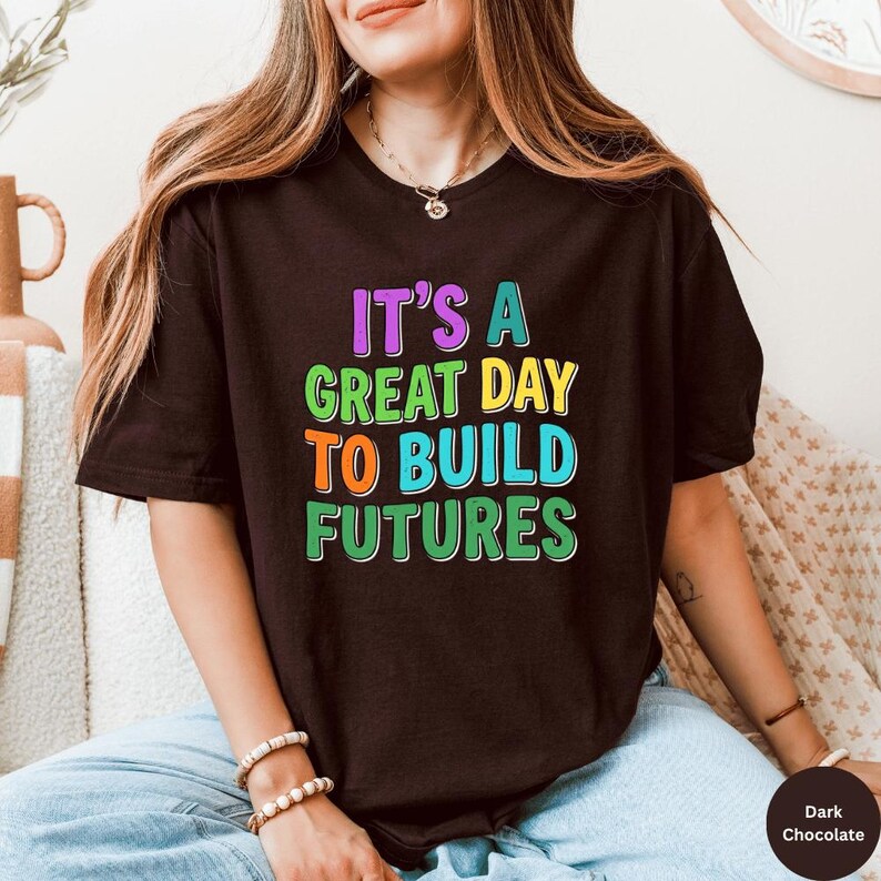 May include: Dark chocolate-colored t-shirt with the text "IT'S A GREAT DAY TO BUILD FUTURES" in colorful, bold letters. The shirt is a short-sleeved crew neck style. The text is in purple, green, orange, and blue.