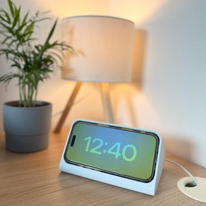 Può includere: Un supporto di ricarica per telefono grigio chiaro con un telefono che mostra l'ora 12:40. Il supporto è su una superficie di legno accanto a una piccola pianta in vaso e una lampada con un paralume beige.