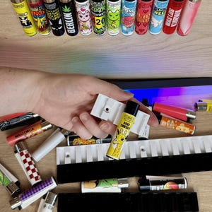 Può includere: Un porta accendini in plastica bianca con 10 slot per accendini. Il porta accendini è pieno di vari accendini colorati con diversi disegni e testi, tra cui "Deep Throat", "Mallorca", "Highway 20", "Peace Week" e "Was Los Kurva".