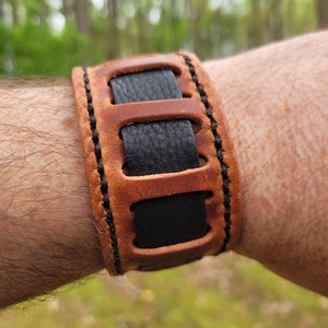 May include: A brown leather bracelet with black leather accents and stitching. The bracelet has a unique design with cutouts and is adjustable.