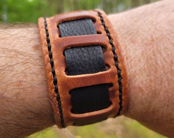 May include: A brown leather bracelet with black leather accents and stitching. The bracelet has a unique design with cutouts and is adjustable.