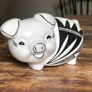 Könnte beinhalten: Ein weißes Keramik-Sparschwein mit schwarzen geometrischen Mustern. Das Sparschwein hat ein lächelndes Gesicht und liegt auf einer Holzoberfläche.