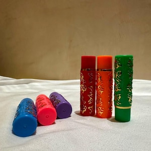 May include: Six cylindrical lipstick tubes in blue, pink, purple, red, orange, and green. Three tubes stand upright while three lie horizontally. Each tube features a textured, raised floral pattern with gold-coloured accents on the exterior surface.
