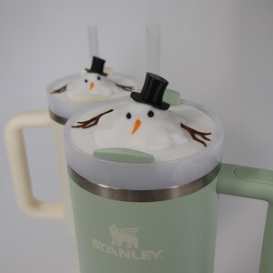 May include: Two green Stanley travel mugs with white lids and snowman toppers. The mugs have black straws and the logo "STANLEY" is printed on the side.