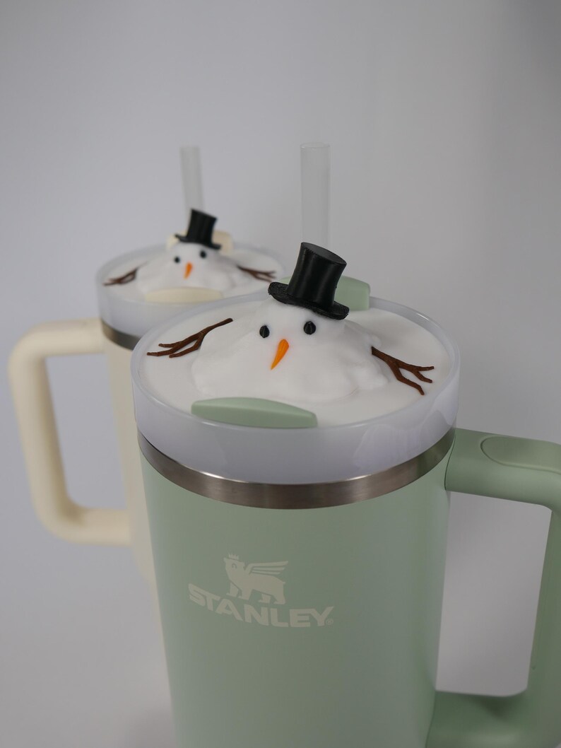 May include: Two green Stanley travel mugs with white lids and snowman toppers. The mugs have black straws and the logo "STANLEY" is printed on the side.