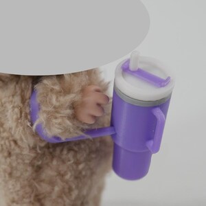 May include: A purple tumbler with a white lid and a handle is held by a furry creature. The tumbler has a straw and a gray band around the middle. The creature's arm and hand are visible, holding the tumbler.