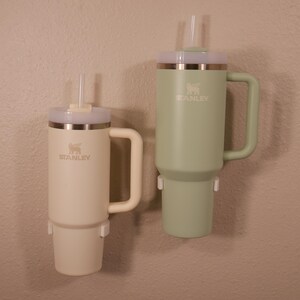 May include: Two stainless steel travel mugs with lids and straws. The mugs are green and beige and have the Stanley logo on them.