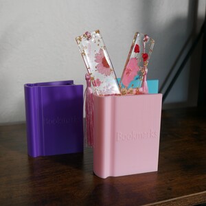 May include: Two pink and clear resin bookmarks with pressed flowers and gold flakes, displayed in a pink and purple book-shaped holder. The holder has the word "Bookmarks" printed on the front.