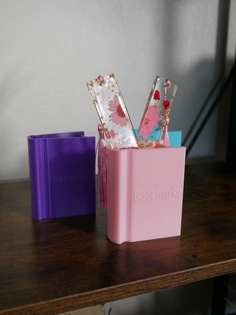 May include: Two pink and clear resin bookmarks with pressed flowers and gold flakes, displayed in a pink and purple book-shaped holder. The holder has the word "Bookmarks" printed on the front.