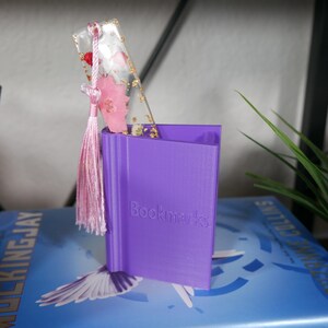 May include: A purple 3D printed bookmark holder with the word "Bookmarks" printed on the front. A clear bookmark with pink flowers and gold flakes is in the holder. The bookmark has a pink tassel.