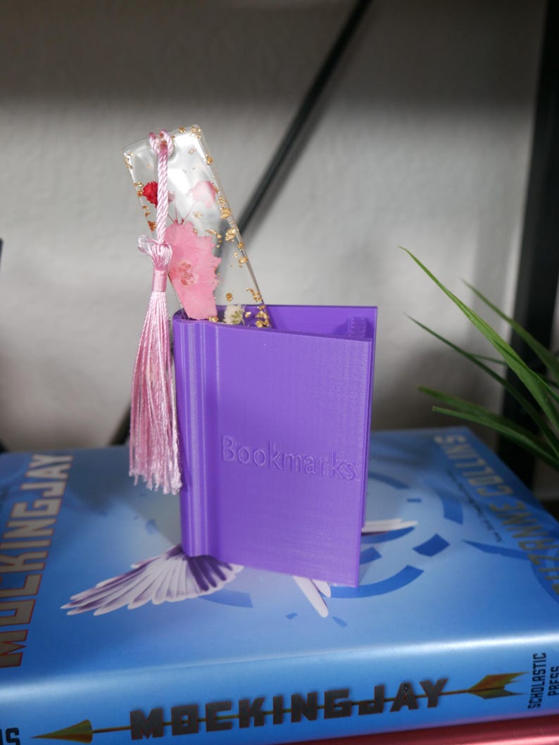 May include: A purple 3D printed bookmark holder with the word "Bookmarks" printed on the front. A clear bookmark with pink flowers and gold flakes is in the holder. The bookmark has a pink tassel.