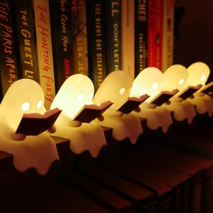 May include: A row of six small, white, ghost-shaped LED lamps, each holding a dark-colored book. The ghosts emit a warm, soft glow from their eyes and bodies, positioned along the edge of a shelf in front of a collection of various books.