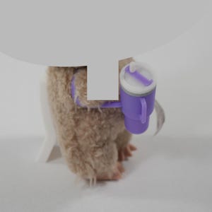 May include: A plush, light brown toy animal with a purple harness and a small purple and white cup attached. The cup has a handle and a lid with a straw. The toy is on a white surface.