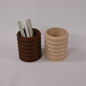 May include: Two stacked ring-style pencil holders, one brown and one beige. The brown holder has three white pens inside.