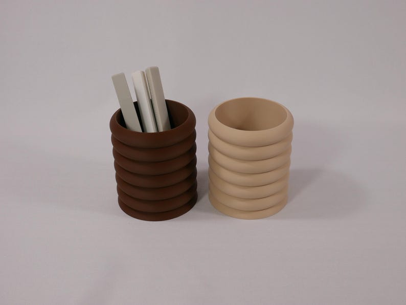 May include: Two stacked ring-style pencil holders, one brown and one beige. The brown holder has three white pens inside.