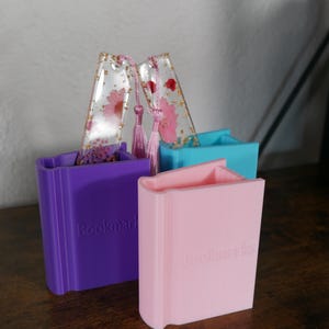 May include: Two book-shaped silicone molds for making resin bookmarks. The molds are purple and pink and have the word "Bookmarks" embossed on the front.