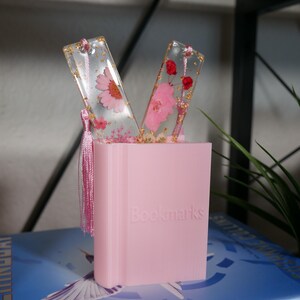 May include: Two pink and gold bookmarks with pressed flowers and gold leaf inside. The bookmarks are displayed in a pink holder that says "Bookmarks".