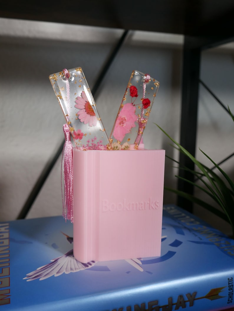 May include: Two pink and gold bookmarks with pressed flowers and gold leaf inside. The bookmarks are displayed in a pink holder that says "Bookmarks".