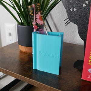 May include: A turquoise blue bookend with the word "Bookmarks" printed on the front. A clear bookmark with pressed flowers and gold leaf is in the bookend.