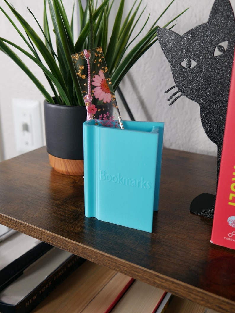 May include: A turquoise blue bookend with the word "Bookmarks" printed on the front. A clear bookmark with pressed flowers and gold leaf is in the bookend.