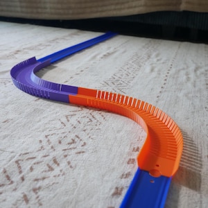 Puede incluir: Un segmento de pista de carreras de juguete con un camino sinuoso construido con piezas de plástico interconectadas. La pista incluye una sección recta larga de color azul, una sección curva morada con paredes laterales acanaladas y una sección curva naranja con costillas.