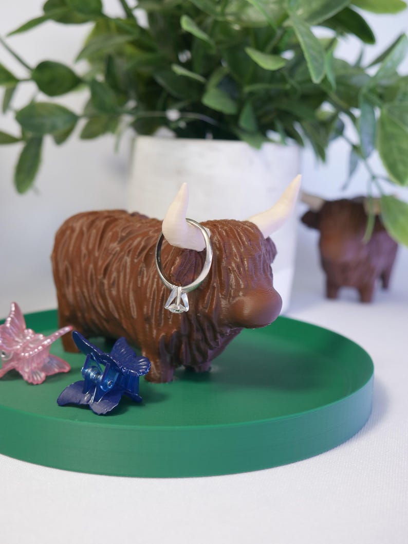 May include: A brown Highland cow figurine with white horns and a ring on its nose, sitting on a green circular tray with two blue butterfly hair clips and a pink butterfly hair clip.
