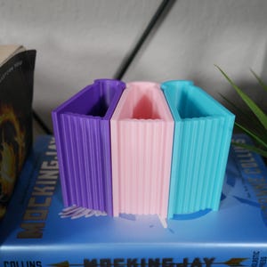 May include: Three bookends shaped like books, in purple, pink, and blue, sitting on top of a blue book with the title "Mockingjay" on the spine. The bookends are made of a textured plastic material.