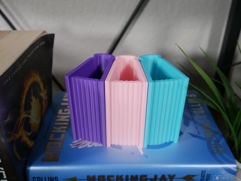 May include: Three bookends shaped like books, in purple, pink, and blue, sitting on top of a blue book with the title "Mockingjay" on the spine. The bookends are made of a textured plastic material.