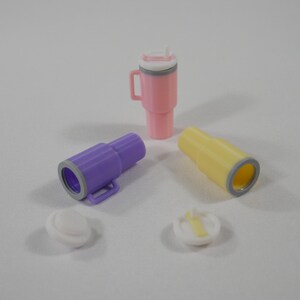 May include: A collection of colorful, miniature plastic drinkware, including a pink tumbler with a handle and straw, two small purple and yellow tubes, and two white lids. The items are arranged on a white surface.